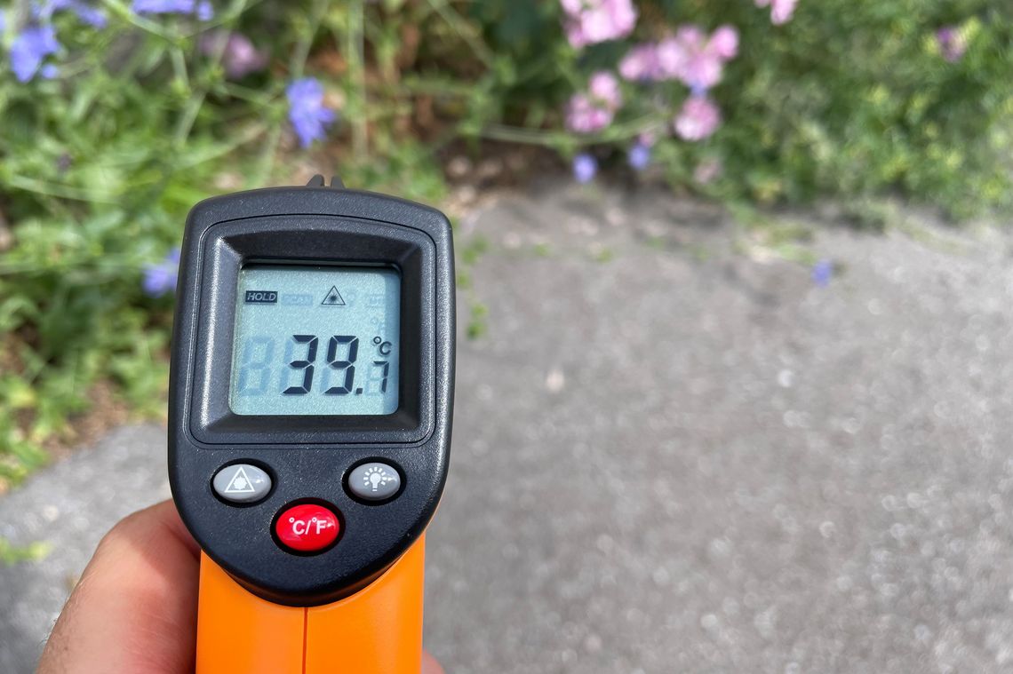 Hand hält Temperaturmessgerät über Asphalt, Anzeige zeigt 39,7 Grad.